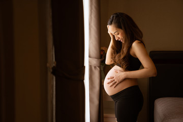 Close-up of a pregnant woman touching her belly on the foreground in emotional smile happy by asian woman with stand relax in bedroom her