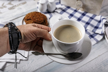Mans hand holding a white cup of coffee. Against the background of a wooden table with cream, sugar bowl and oatmeal cookies. Copy space. Horizontal shot