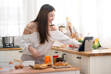 Young Asian woman prepare for breakfast in kitchen, using digital tablet computer
