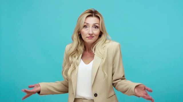 Confused adult woman in business jacket shrugging shoulders, puzzled by information, uncertain. Indoor studio shot isolated on blue background