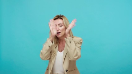 Scared terrified adult woman in business suit closing eyes and stretching hands out trying to protect herself, victim afraid of abusers. Indoor studio shot isolated on blue background