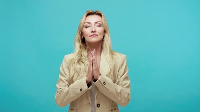 Relaxed and calm middle aged businesswoman pressing hands together with smile on her peaceful face, making namaste gesture, practicing yoga to calm. Indoor studio shot isolated on blue background