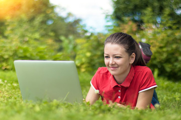 happy girl with laptop lying on the grass in the park on sunny day. young woman working remotely at the computer. female leisure with gadgets. student studying in nature