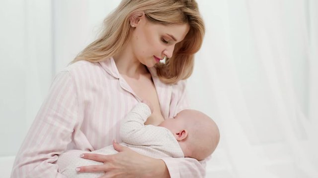A young caucasian mother breastfeeds a newborn baby sitting on a bed in a light bedroom.