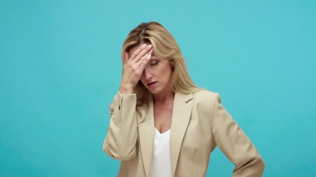 Exhausted tired adult woman with blond hair doing facepalm gesture, shocked and depressed with stupid situation, ashamed with mistake, loser. Indoor studio shot isolated on blue background
