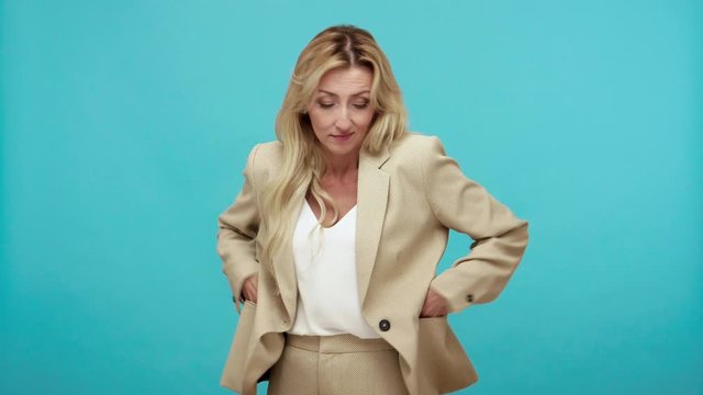 Poor jobless adult woman with blond hair has no money showing empty pockets of her stylish jacket, dismissal, crisis, monetary reform. Indoor studio shot isolated on blue background