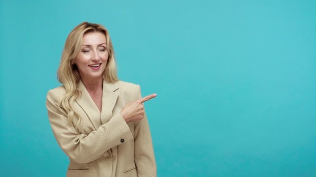Happy surprised blond woman in suit pointing finger away showing free space for advertising, satisfied with advertisement banner, service. Indoor studio shot isolated on blue background