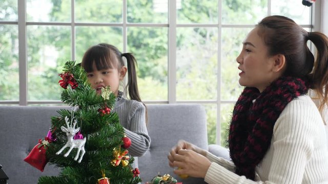 Handheld, Normal view, medium shot of happy asian mother and young daughter, in sweather, decorating christmas tree in the living room