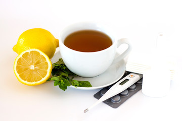 white cup with tea next to lemon, mint, thermometer and pills in a blister on a white background. Seasonal winter, autumn illnesses, colds or flu. Home alternative treatment concept.