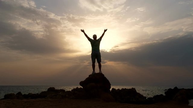 dark male silhouette, man stands on rocks in front of the ocean or sea, with his arms outstretched and hands up at dawn, sunrise. concept of human strength and faith