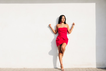 portrait of elegant latin young girl with long brunette hair and red dress on white wall with strong shadow, looking at camera