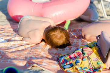 A small child with red hair sleeps on the beach. The child is curled up in a ball and sleeps under the rays of the sun.