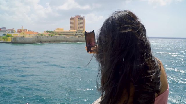 Young girl makes photo pictures to smartphone on promenade with ocean bay and city buildings view  in Willemstad Curacao