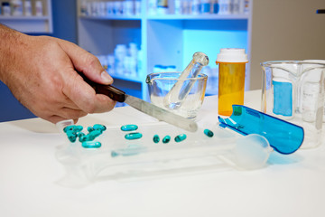 An image of a hand using a pill counting tray with other pharmacy equipment on a countertop and pill bottles in shelves in the backgound lit with blue light