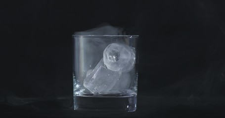Ice cubes melt in the alcohol, whiskey glass and smoke is floating against a black background