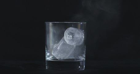 Ice cubes melt in the alcohol, whiskey glass and smoke is floating against a black background