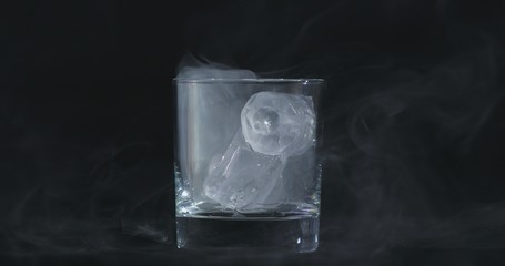 Ice cubes melt in the alcohol, whiskey glass and smoke is floating against a black background