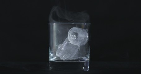 Ice cubes melt in the alcohol, whiskey glass and smoke is floating against a black background