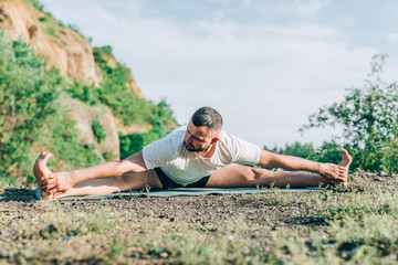 Fototapeta na wymiar Fitness, people and healthy lifestyle concept. Bearded man doing yoga side crane pose on mat outdoors over quarry lake background. International Day of Yoga.