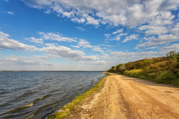 Fototapeta premium The road along the shore of the lake, river, against a blue sky with belvmi clouds