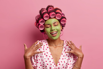 Portrait of pleased young African American woman stands with closed eyes, smiles gently, imagines something nice, applies green face mask, hair curlers, stands indoor, pink background. Domestic style