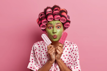 Portrait of good looking woman keeps lips rounded, applies green facial mask, hair rollers, holds two delicious ice creams, dressed in silk gown, poses over pink background. Skin care concept