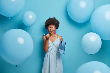 Fototapeta premium Stylish African American woman holds mobile phone near mouth, makes voice call, poses in stylish dress with high heel shoes, dresses for holiday event, stands indoor near inflated helium balloons.