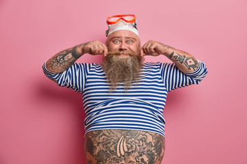 Joyful stout man curls mustache, has thick beard, cheerful face expression, wears swimming hat and...
