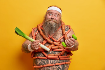 Funny fatso guy poses with sport equipment and green leek, wrapped by sausages, tries to eat...