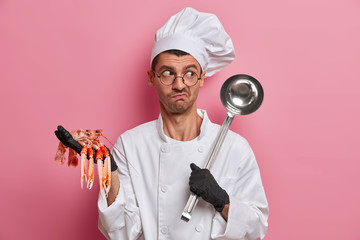 Puzzled chef holds raw crayfish and ladle, going to prepare soup from sea food, wears uniform, hat,...