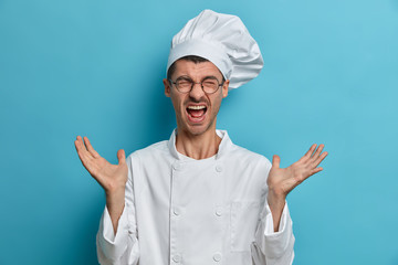 Busy emotional chef raises hands and screams loudly, has much work at kitchen, wears round...