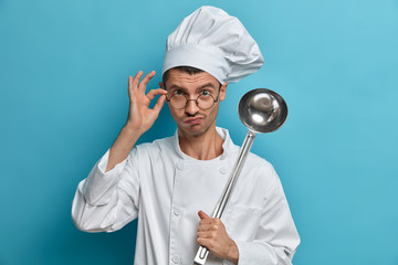 Chef cook poses at commercial kitchen, looks seriously through glasses, holds ladle, prepares meal, ready to cook dish, listens advice, learns restaurant menu, isolated on blue wall. Culinary concept