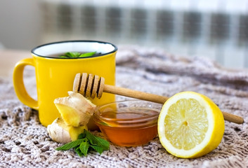 A yellow cup of tea on a beige blanket next to lemon, ginger, a spoonful of honey and green mint. Seasonal illnesses and colds. Home treatment with hot drinks. side view