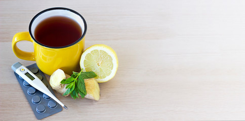 Top view A yellow cup of tea standing on a light table next to lemon, ginger, thermometer and pills. Seasonal illnesses, flu and colds. Space for text