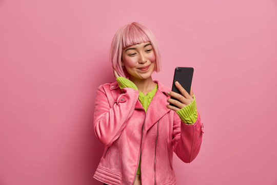 Portrait of charming Asian girl takes selfie on smartphone, makes photo of herself for fashion blog, has pink hairstyle, smiles positively, rosy background, enjoys pleasant online communication