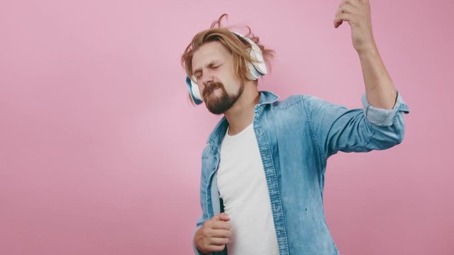 Mature man with trendy beard and haircut enjoying music in modern headphones over pink background. Happy guy singing and dancing in studio.