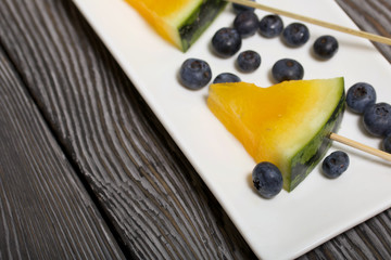 Pieces of yellow watermelon on sticks. Lie on a plate. Nearby are blueberries. Plate on painted pine boards.
