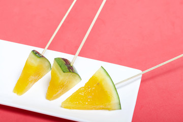 Pieces of yellow watermelon on sticks. Lie on a plate. A plate on a coral colored surface.