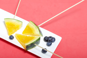 Pieces of yellow watermelon on sticks. Lie on a plate. Nearby are blueberries. A plate on a coral colored surface.