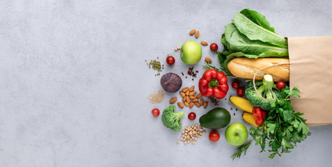 Vegetarian grocery shopping set. Greens, peppers, avocado, broccoli, apple, salad and different types of cereals come out of a paper bag on a gray background.Banner,copy space.