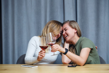 two girls of European appearance with blond hair are sitting at the table, drinking wine and laughing, relaxing at home, alcohol