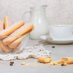 Italian savoiardi or ladyfingers biscuits on a plate and pieces of cookies on the table, a white cup of coffee with a spoon and a jug of milk in the background. Selective focus.