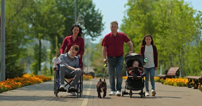 Disabled guy is being driven by his mother in a wheelchair, a family walk. Family with a teenager in a wheelchair, a toddler in a stroller and a dog are walking in the park. 4k, 10bit, ProRes
