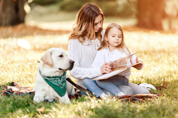 Happy beautiful mother and daughter with dog labrador is reads book sitting in the park.