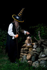 A woman in a black conical hat and a black Halloween costume with a lamp in her hands and a metal hook at night.