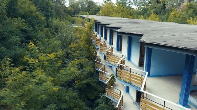 Abandoned hotel or motel among a dense green forest. Deserted building with broken windows and balconies. Aerial side view