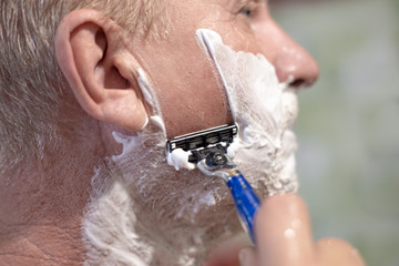 Close up of an adult man with shaving foam on his face, shaving with a reusable razor. Facial care