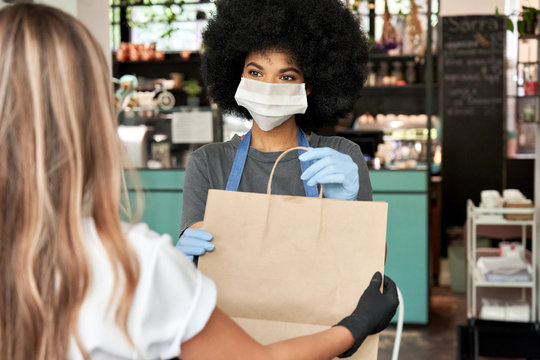 African American female cafe worker wears face mask and gloves giving takeaway food bag to customer. Mixed race waitress holding takeout order standing in coffee shop restaurant with take away client.
