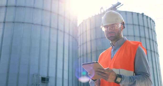 Close up portrait of busy Caucasian man engineer in uniform and helmet in factory yard outdoor and tapping on tablet. Man in goggles standing near big cisterns and typing on device. Factory concept