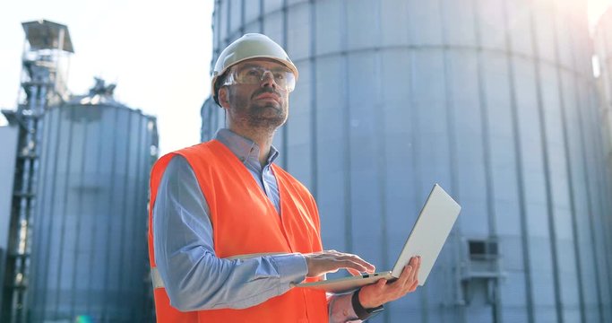 Side view of handsome Caucasian male worker in helmet and goggles typing on laptop while standing at plant territory near big tanks. Portrait of man constructor working on computer outdoor Job concept
